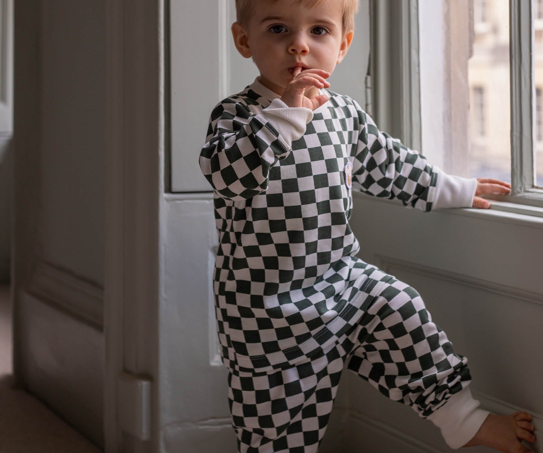 Child wearing a black and white checkered outfit standing by a window.
