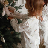 Child in pajamas decorating a Christmas tree with a teddy bear ornament.