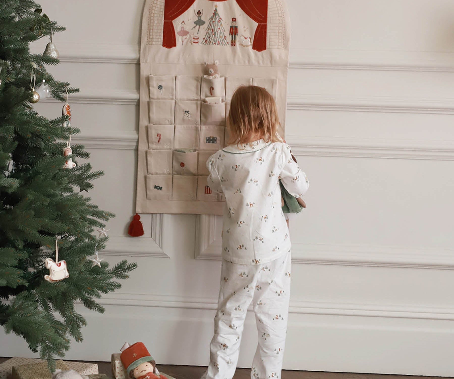 Child in pajamas looking at a Christmas-themed calendar next to a decorated tree.