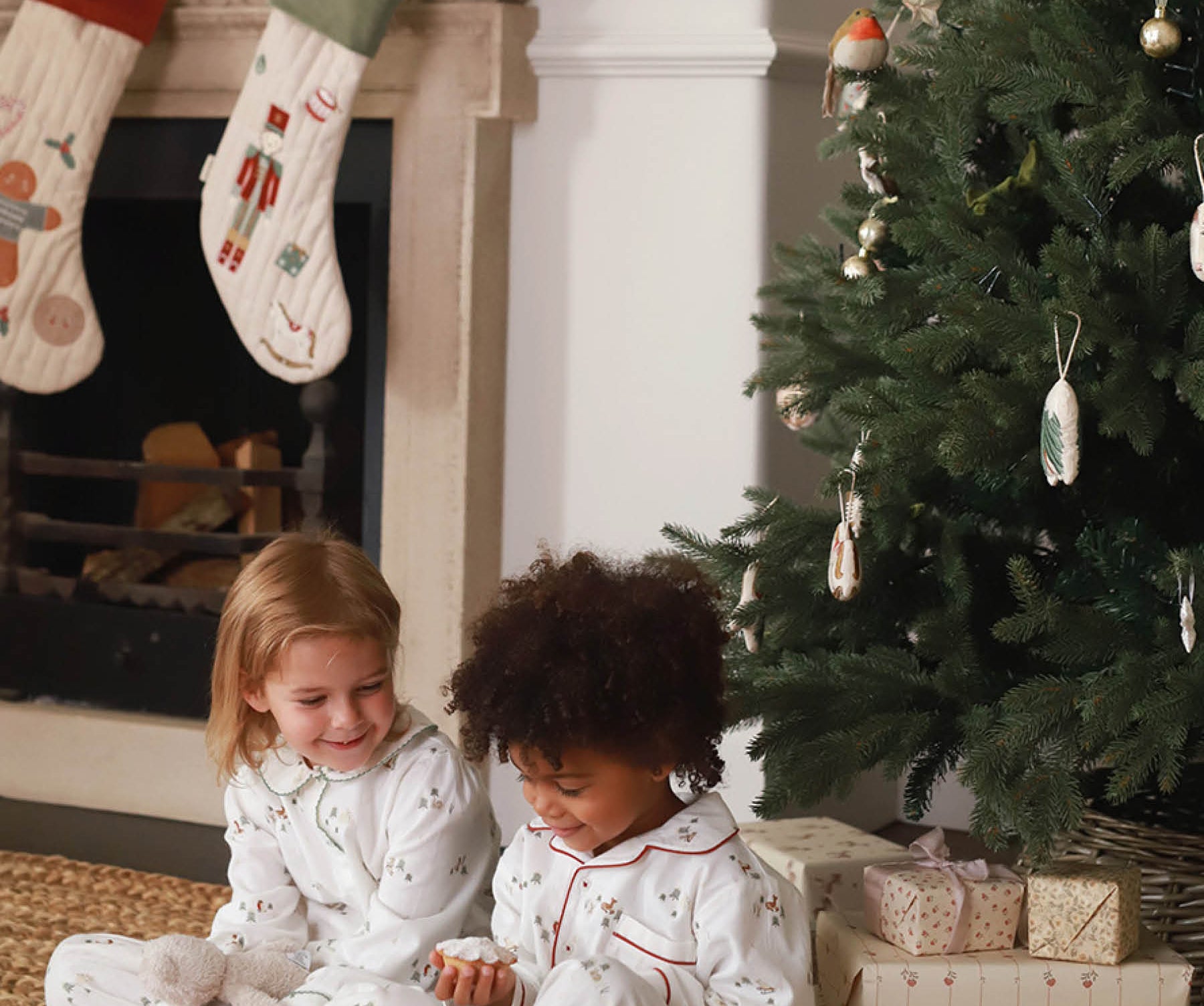 Two children in pajamas sitting by a Christmas tree and fireplace.