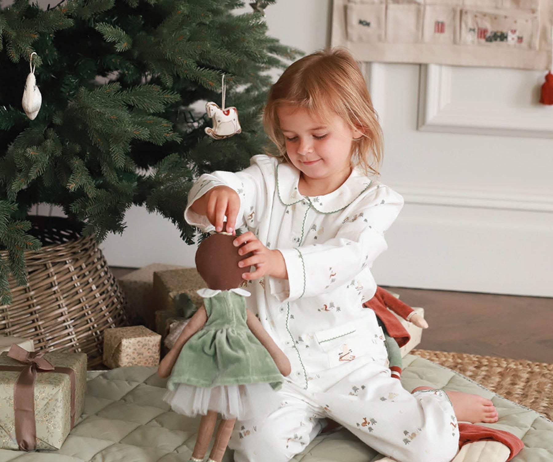 Child in pajamas playing with a doll next to a decorated Christmas tree.