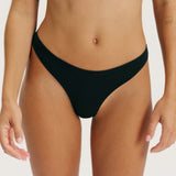 A close-up of a model wearing a solid black bikini bottom against a clean, off-white background.