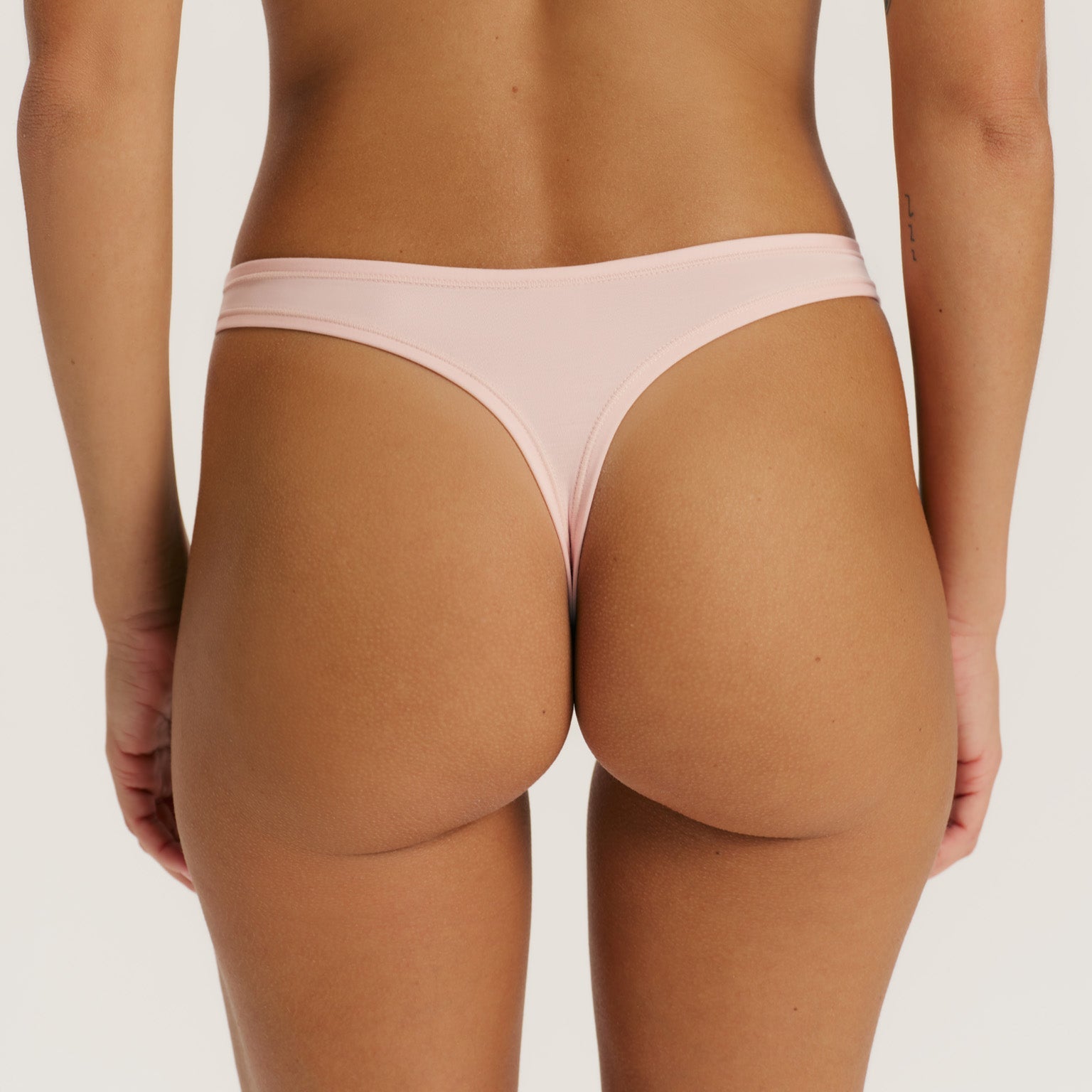 Rear view of a model wearing a light pink thong-style underwear bottom against a plain light gray studio background.