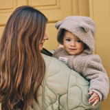ALF Eco Pramsuit in beige worn by a smiling toddler with the bear-ear hood up, held by a woman in a quilted green coat