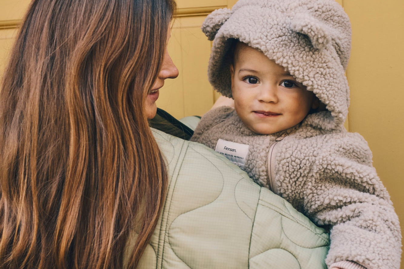 ALF Eco Pramsuit in beige worn by a smiling toddler with the bear-ear hood up, held by a woman in a quilted green coat