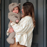 Alf Eco Pramsuit Beige on a baby with the hood up, held in a woman's arms in front of a dark door