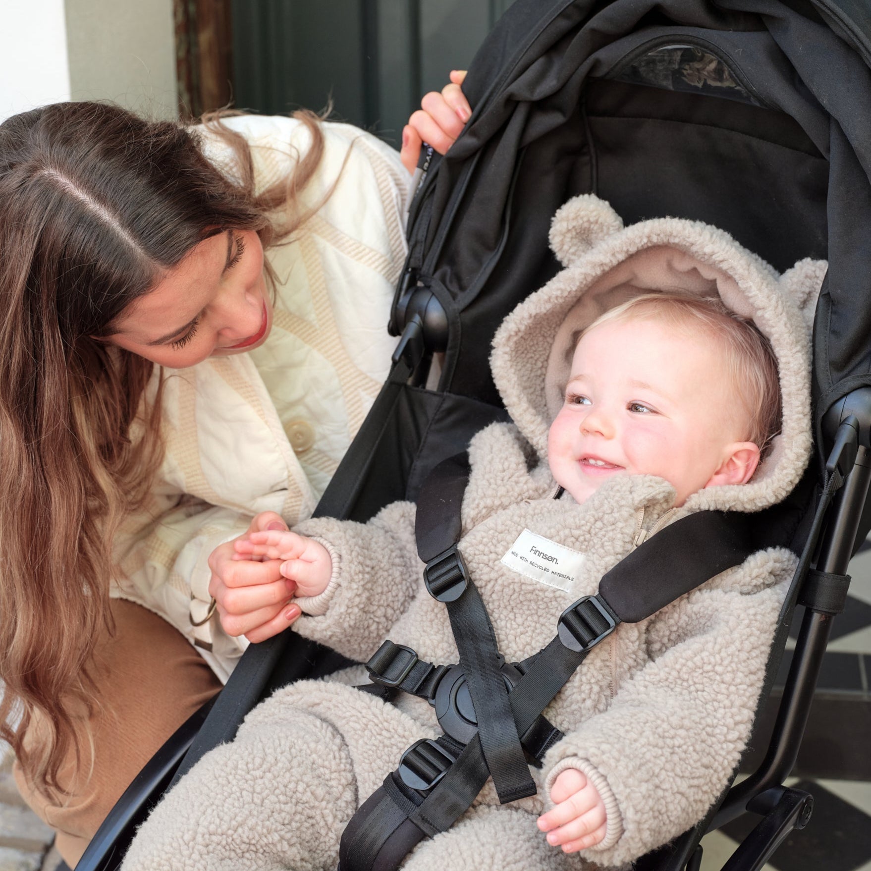ALF Eco Pramsuit in beige worn by a smiling baby in a pushchair while a woman crouches beside them