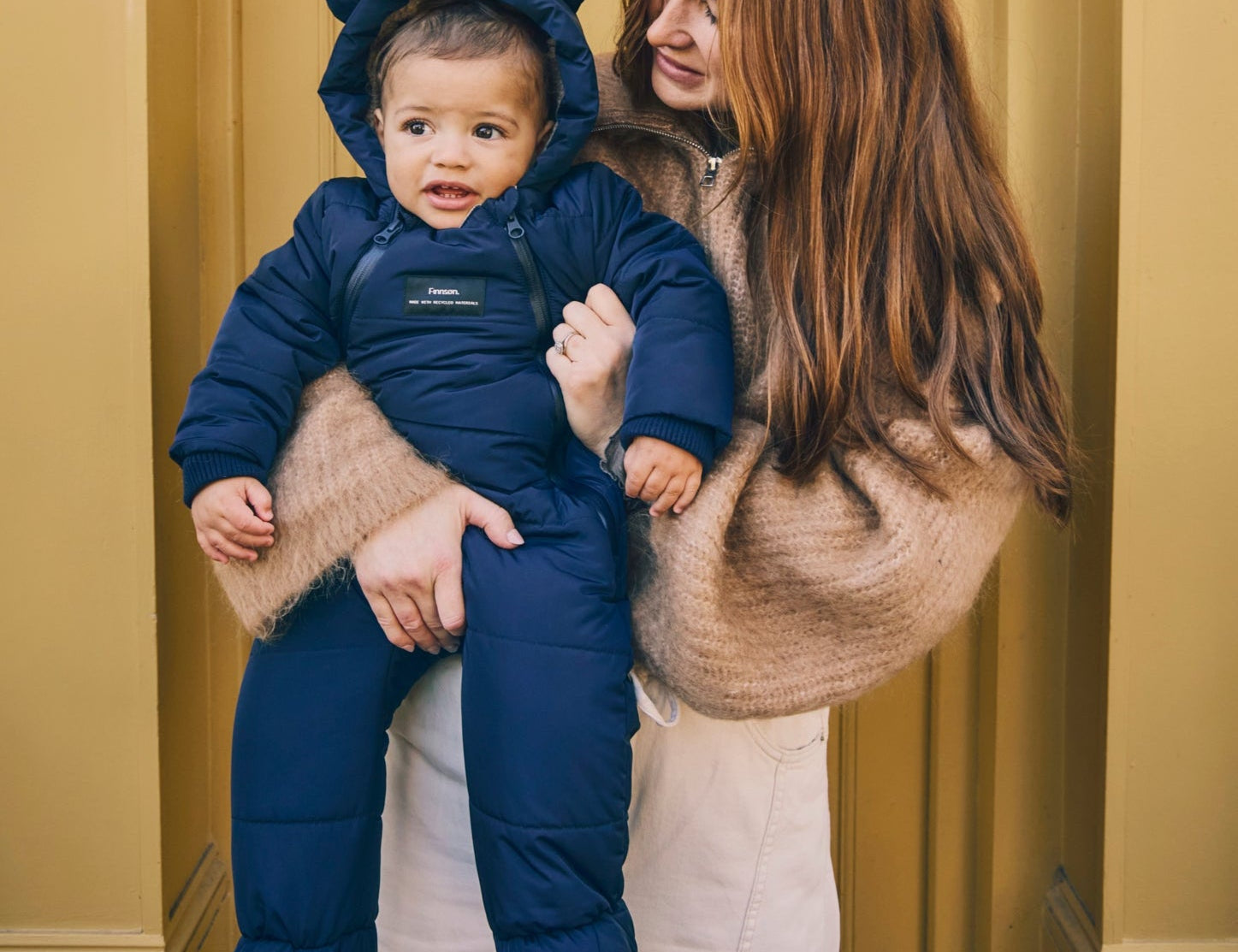 Anders Eco Snowsuit in navy worn by a baby held on the hip of a woman standing in front of a yellow door