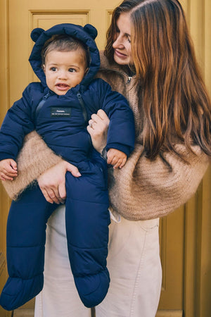 Anders Eco Snowsuit in navy worn by a baby held on the hip of a woman standing in front of a yellow door
