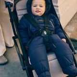 Anders Eco Snowsuit in navy worn by a baby with hood up, strapped into a pushchair seat