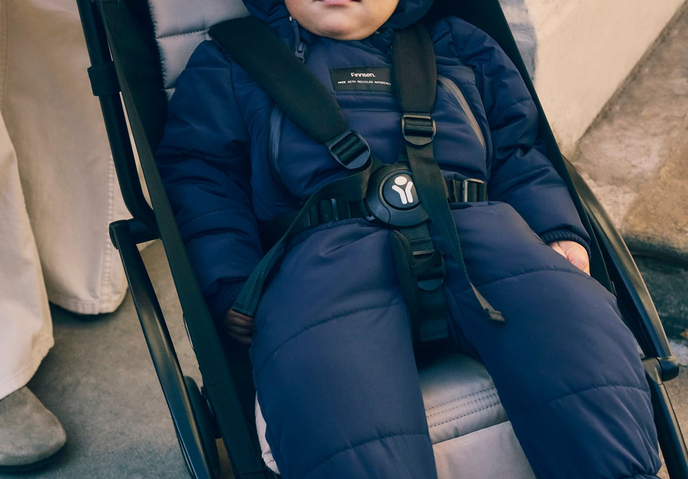 Anders Eco Snowsuit in navy worn by a baby with hood up, strapped into a pushchair seat