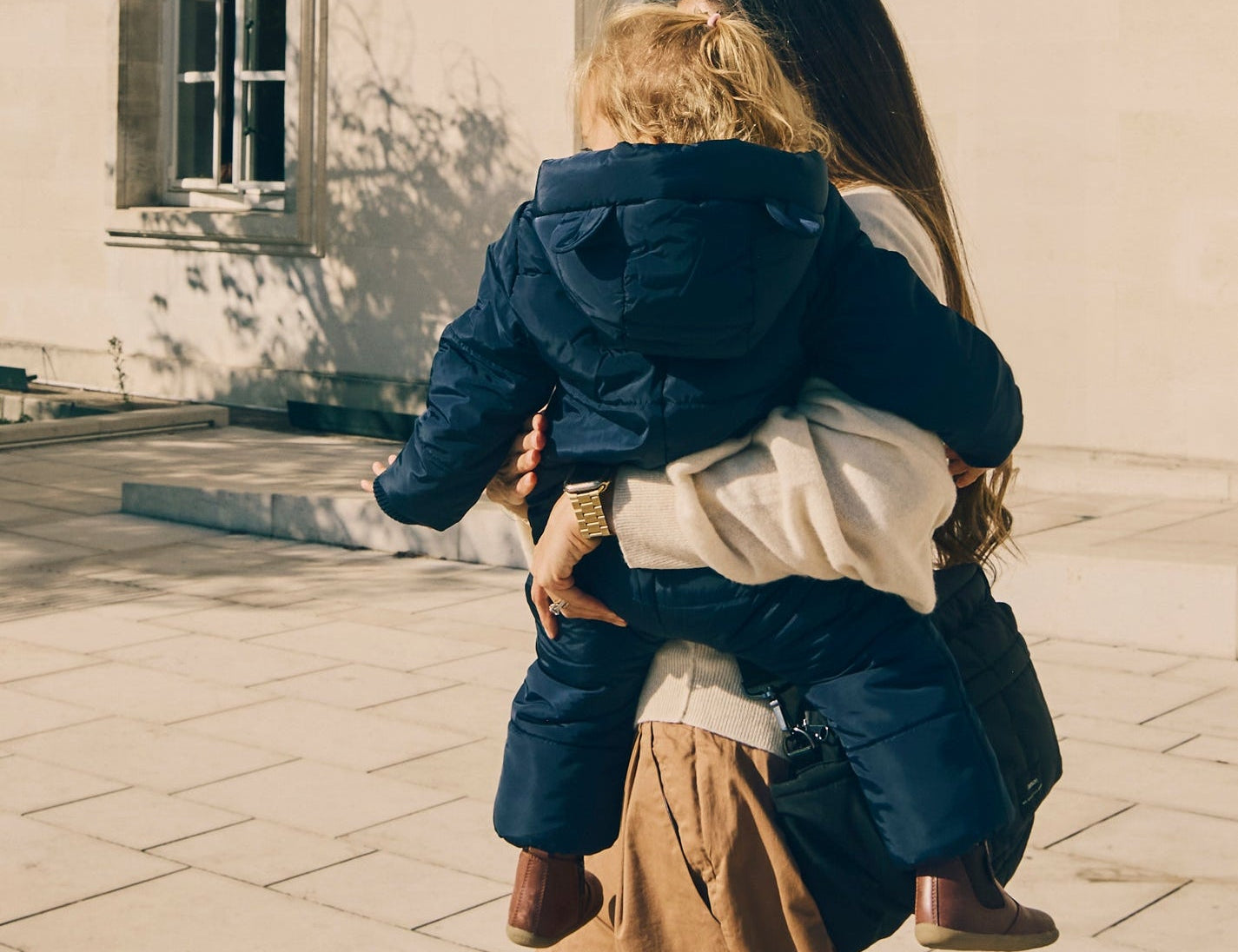 Anders Eco Snowsuit Navy worn by a toddler being carried on a woman's hip, seen from behind outdoors