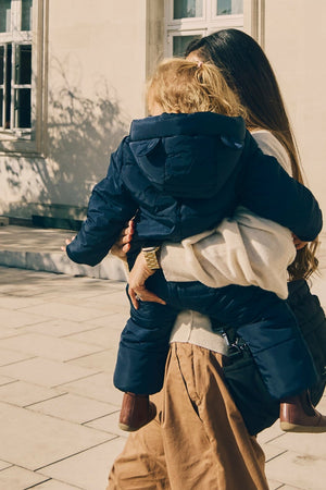 Anders Eco Snowsuit Navy worn by a toddler being carried on a woman's hip, seen from behind outdoors
