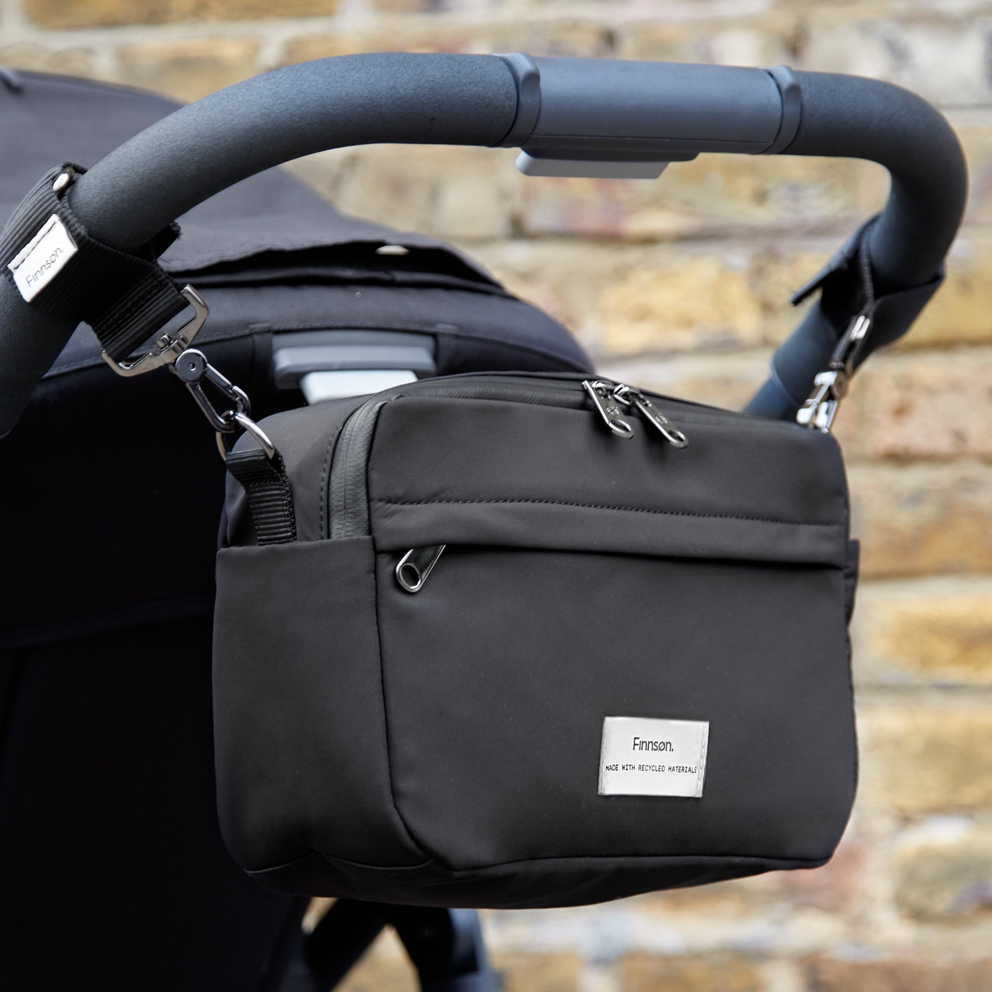Frida Eco Stroller Organiser Black hanging from a pushchair handlebar against a brick wall