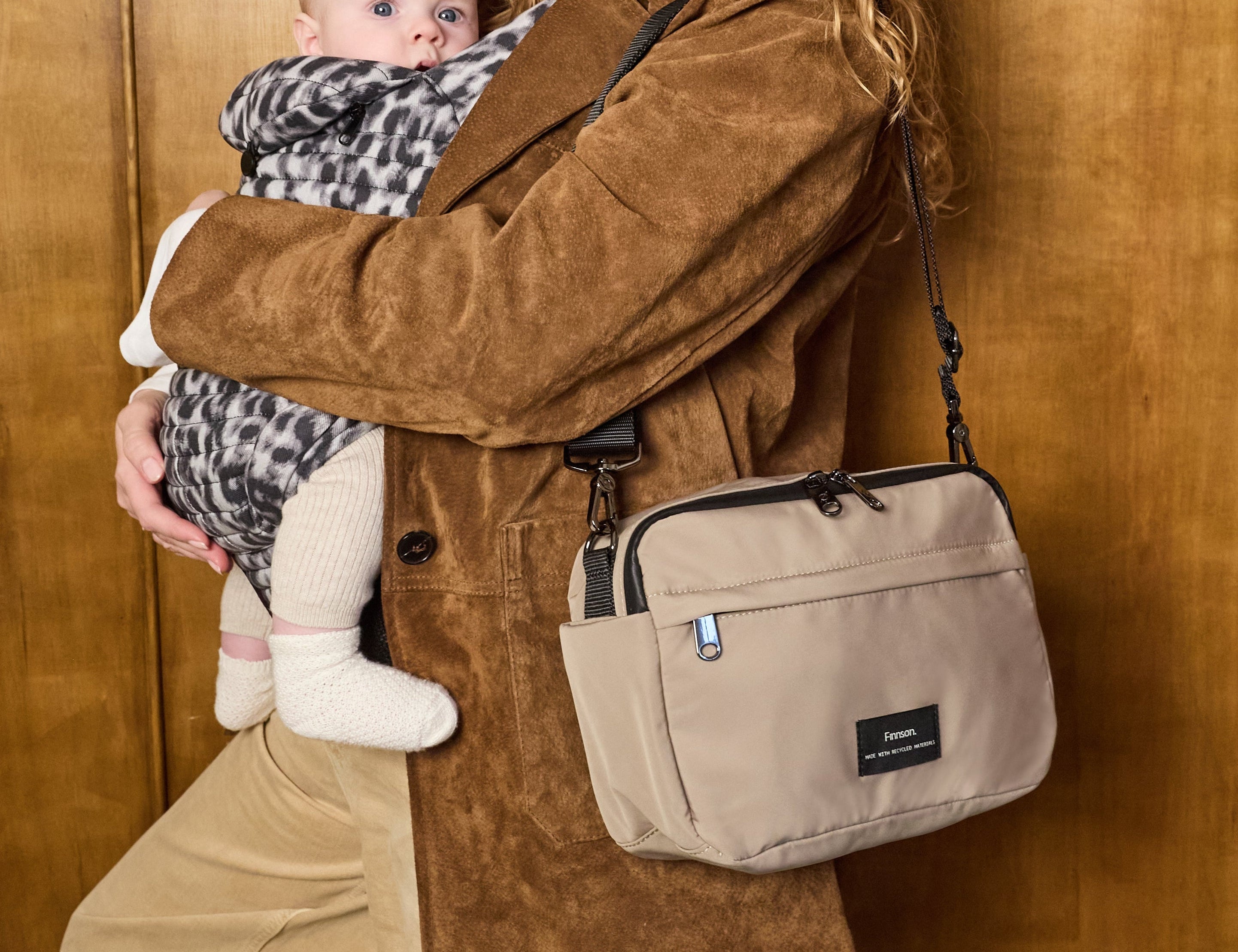Frida Eco Stroller Organiser Taupe worn crossbody by a woman in a suede coat while she holds a baby in a carrier