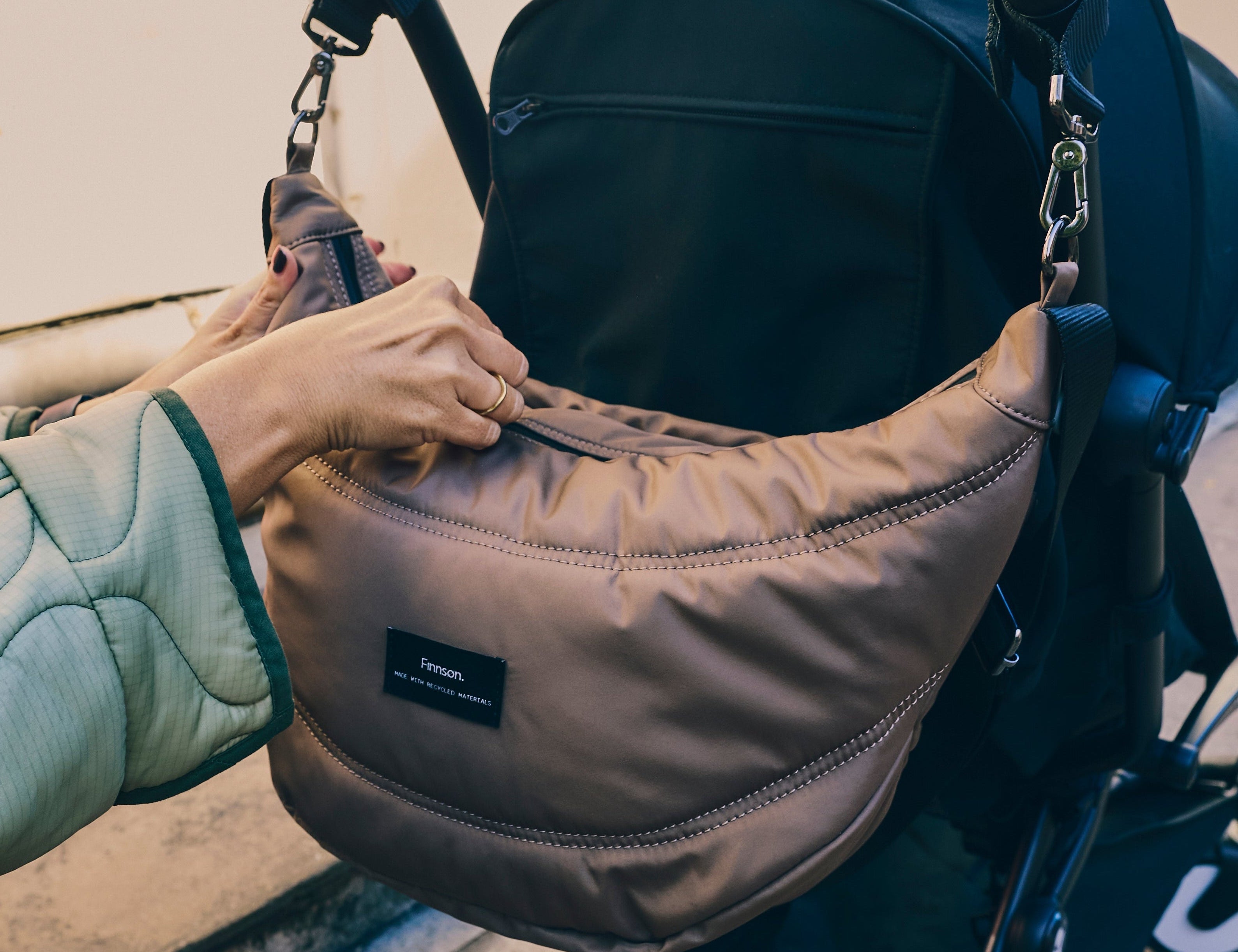Osma stroller organiser in Mocha clipped to a stroller handle while a hand reaches into the top zip