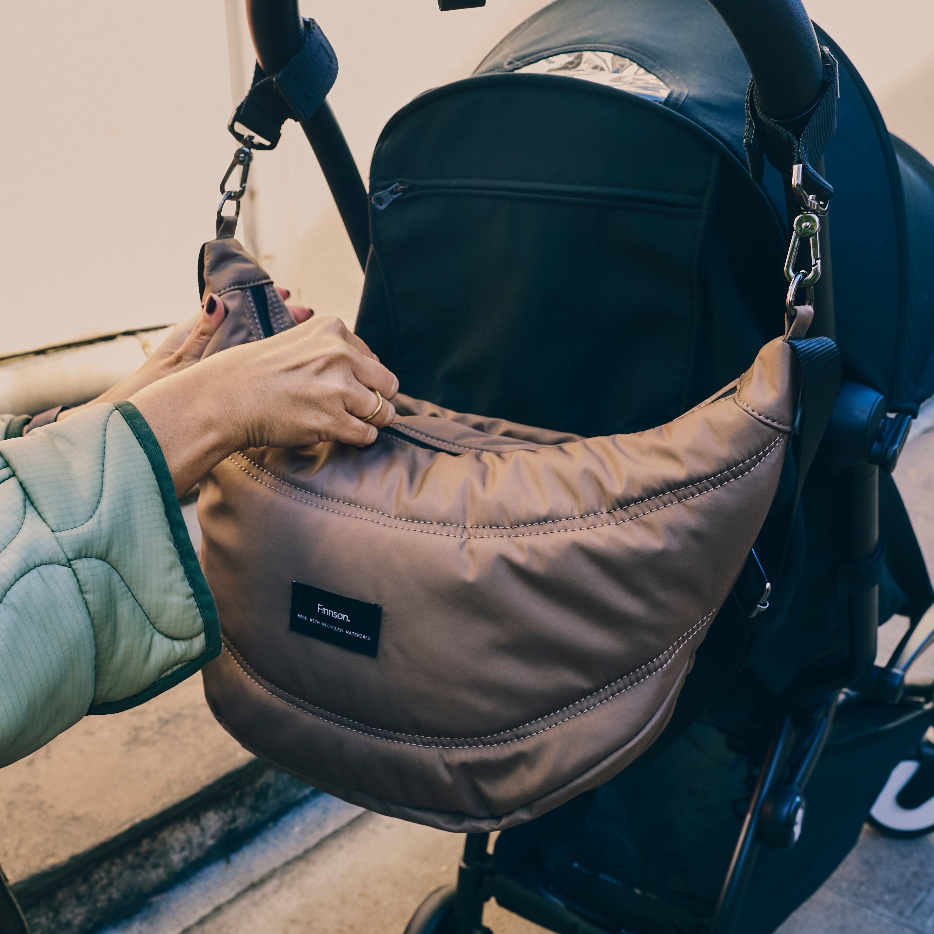 Osma stroller organiser in Mocha clipped to a stroller handle while a hand reaches into the top zip