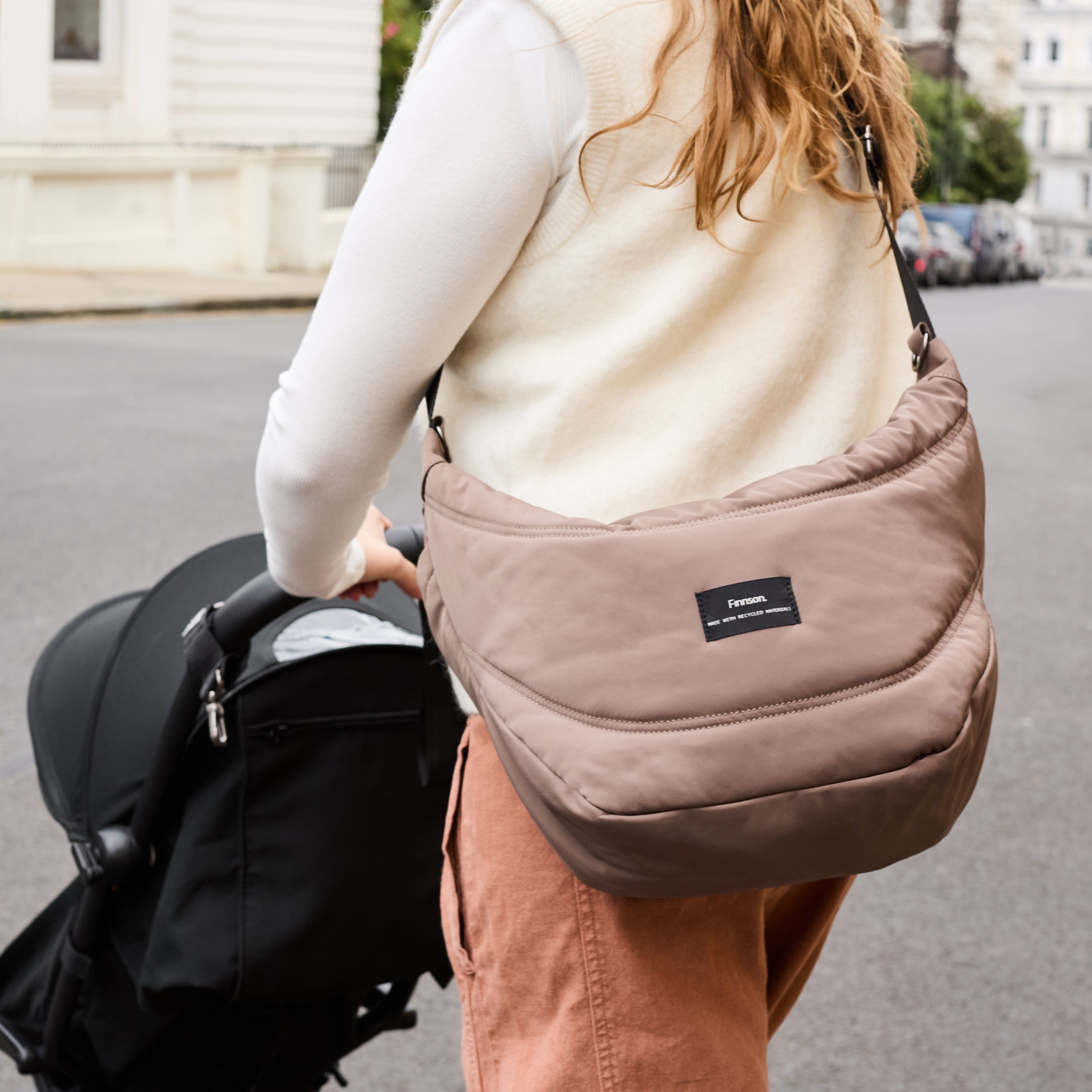 Woman wearing the Osma stroller organiser in Mocha across her body, seen from behind while she pushes a stroller
