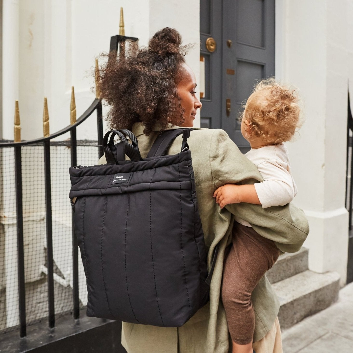 Woman carrying a child on her back with a black backpack, standing outside a building.