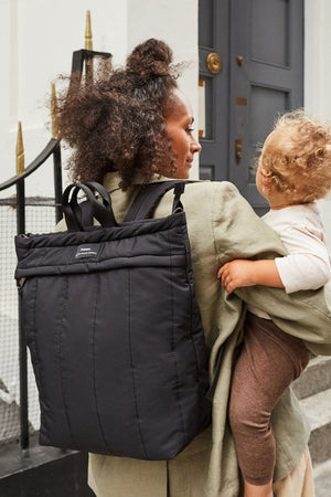 Woman carrying a child on her back with a black backpack, standing outside a building.