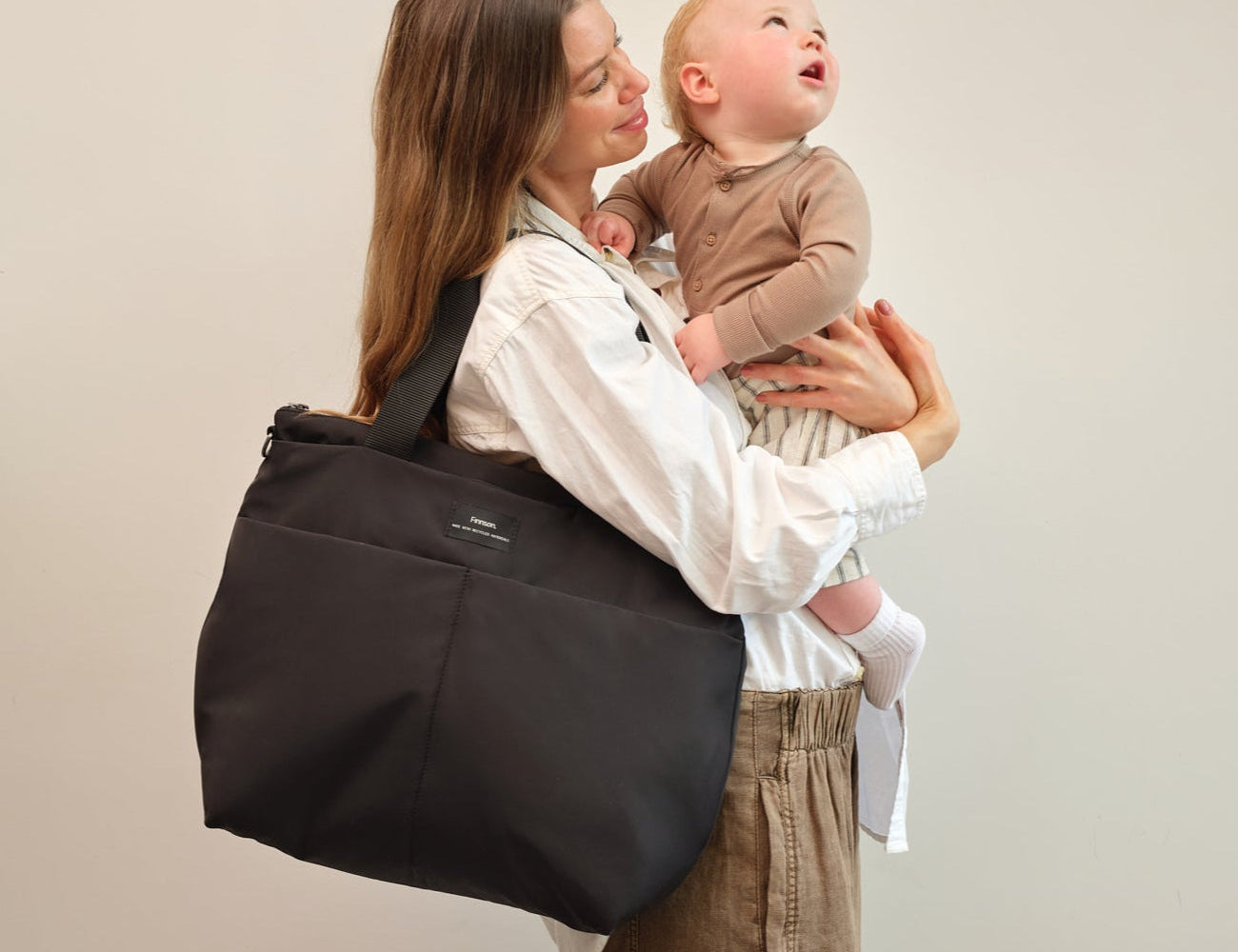 Sigrid Eco Changing Bag black, worn on the shoulder by a woman holding a baby who looks upwards