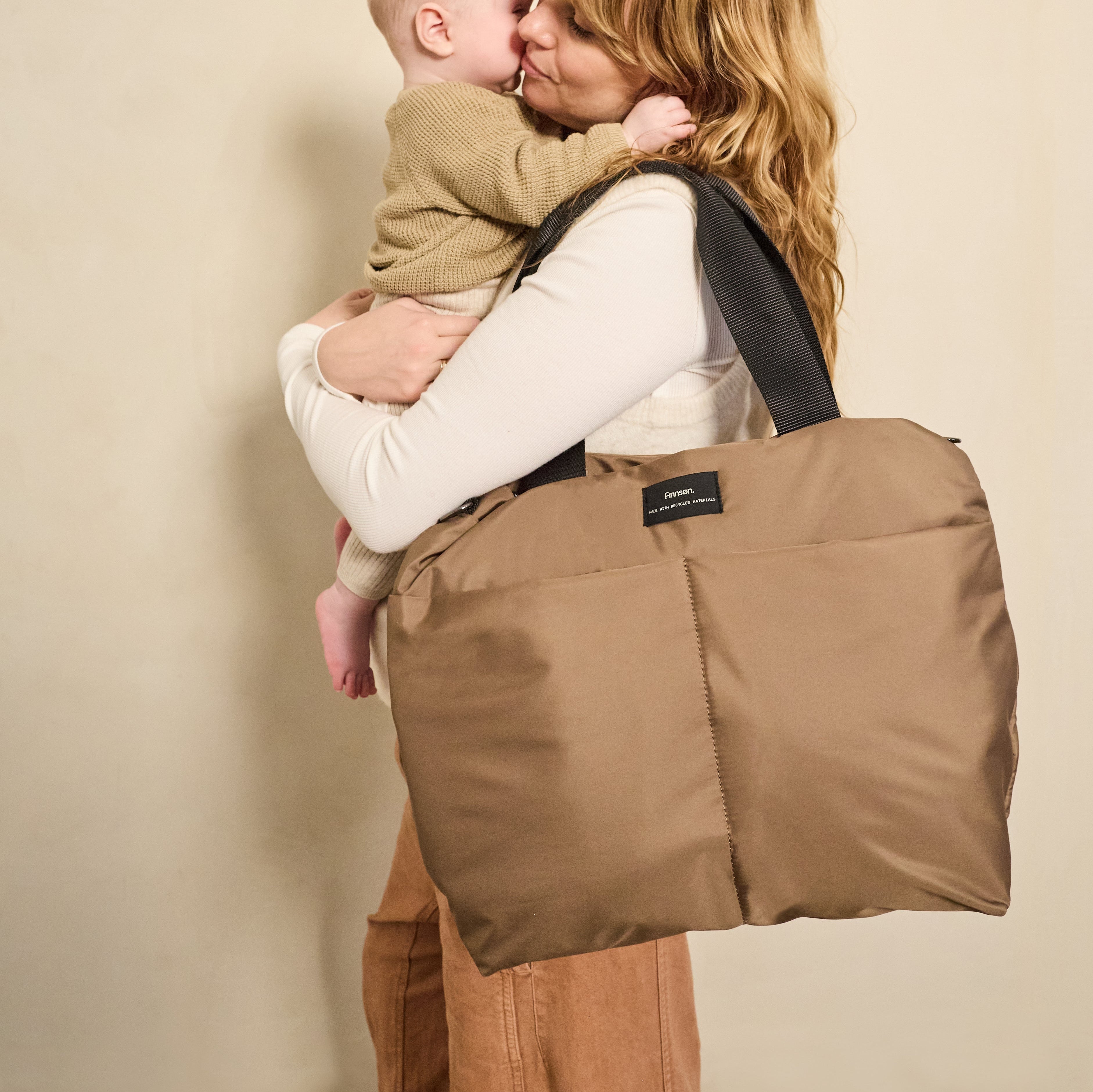 Sigrid Eco Changing Bag in Mocha worn on the shoulder by a woman as she cuddles and kisses a baby