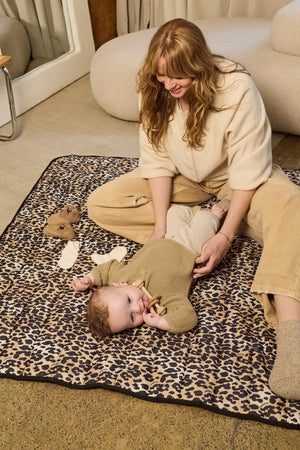 Thoren Eco Playmat in Leopard indoors with a woman sitting beside a baby lying on it holding a wooden toy