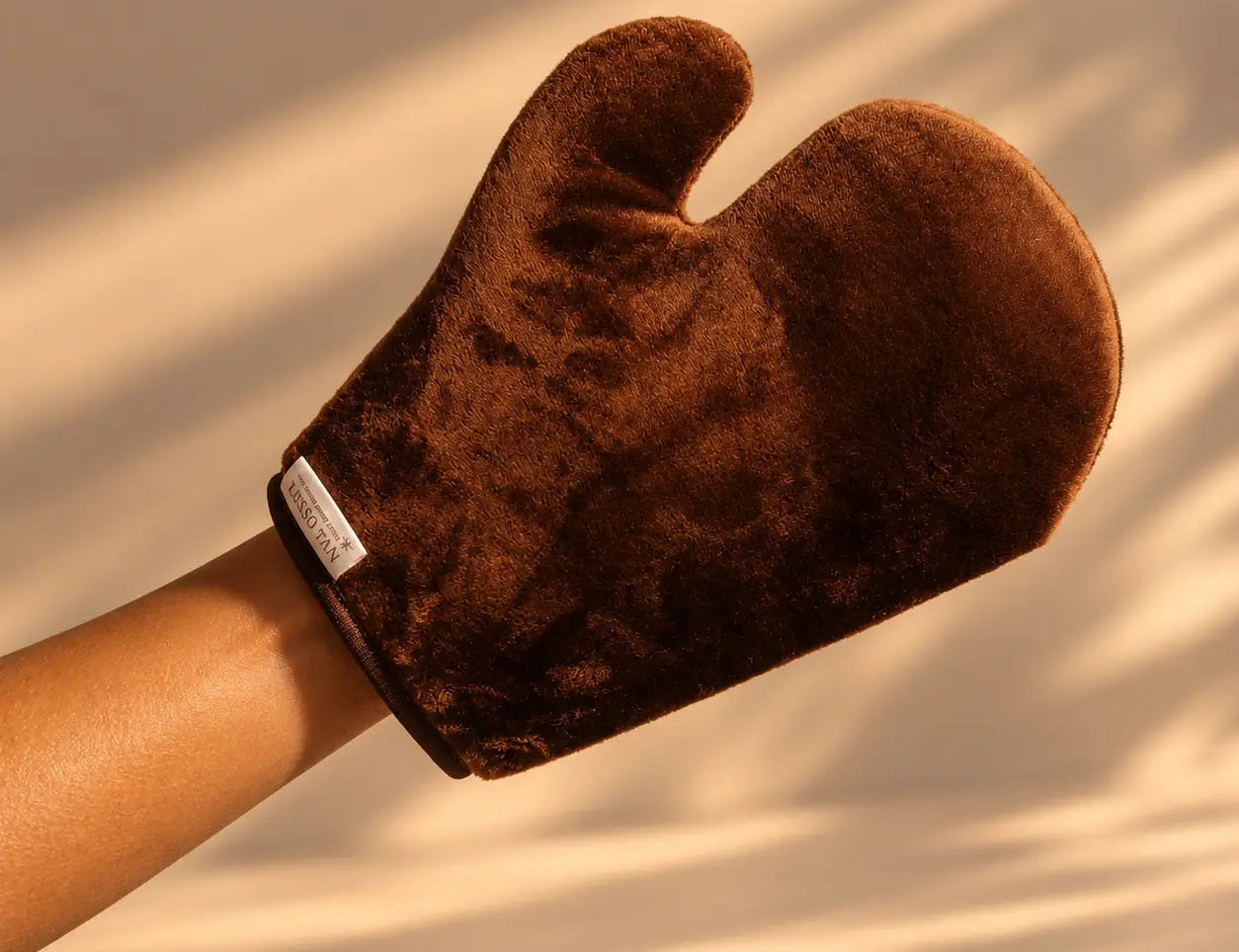 Flawless Tanning Mitt in brown, worn on a raised hand and forearm against a wall with soft sunlight shadows