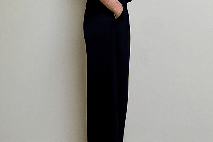 Person wearing a black outfit standing against a white wall on a wooden floor.