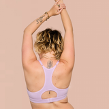 A woman with blonde-highlighted curly hair is shown from behind wearing a light lavender sports bra with a large open back cutout. She has her arms raised overhead, standing against a solid pale peach background.