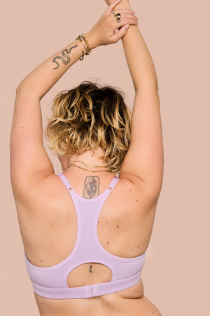 A woman with blonde-highlighted curly hair is shown from behind wearing a light lavender sports bra with a large open back cutout. She has her arms raised overhead, standing against a solid pale peach background.