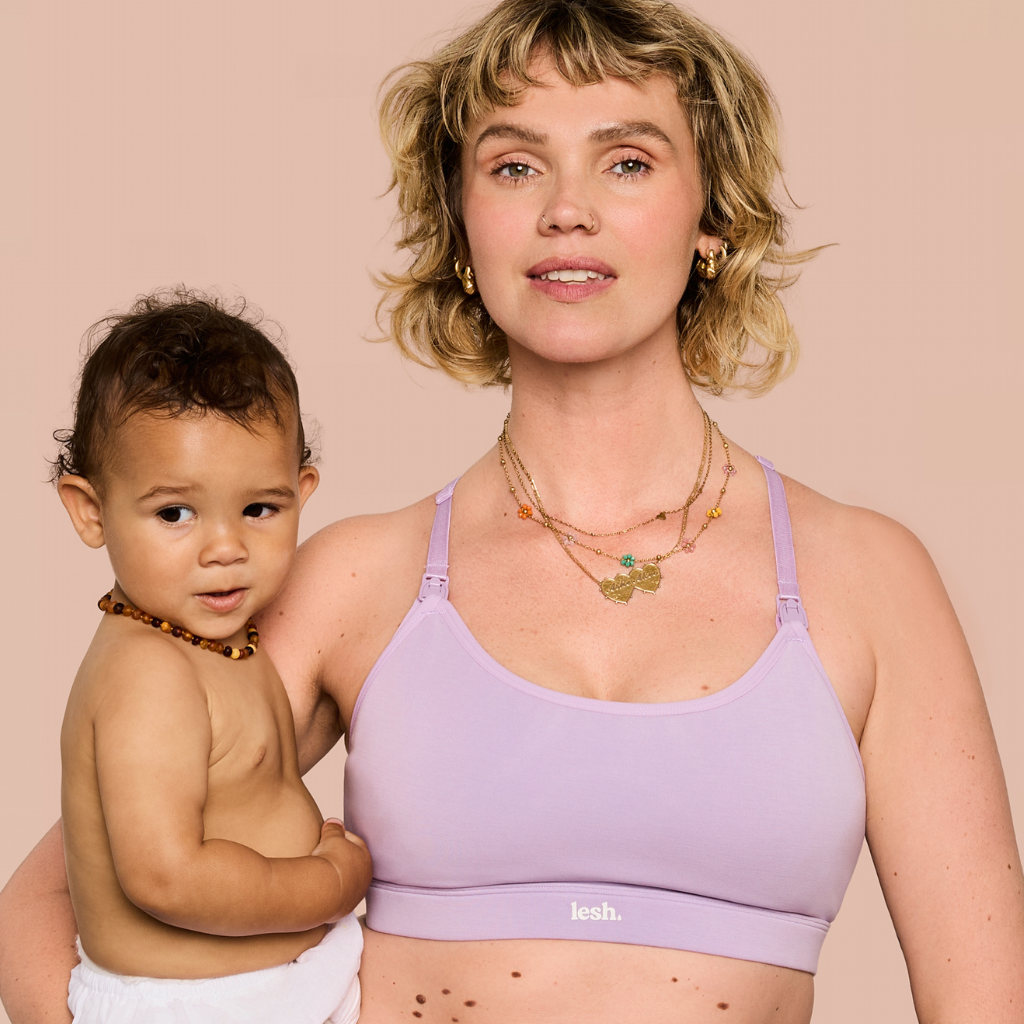 A woman holding a baby wearing a light purple/lavender nursing bra with the brand name 'leeh.' visible on the band, against a solid pale pink background.