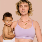 A woman holding a baby wearing a light purple/lavender nursing bra with the brand name 'leeh.' visible on the band, against a solid pale pink background.