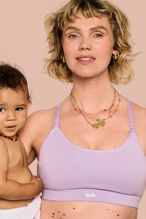 A woman holding a baby wearing a light purple/lavender nursing bra with the brand name 'leeh.' visible on the band, against a solid pale pink background.