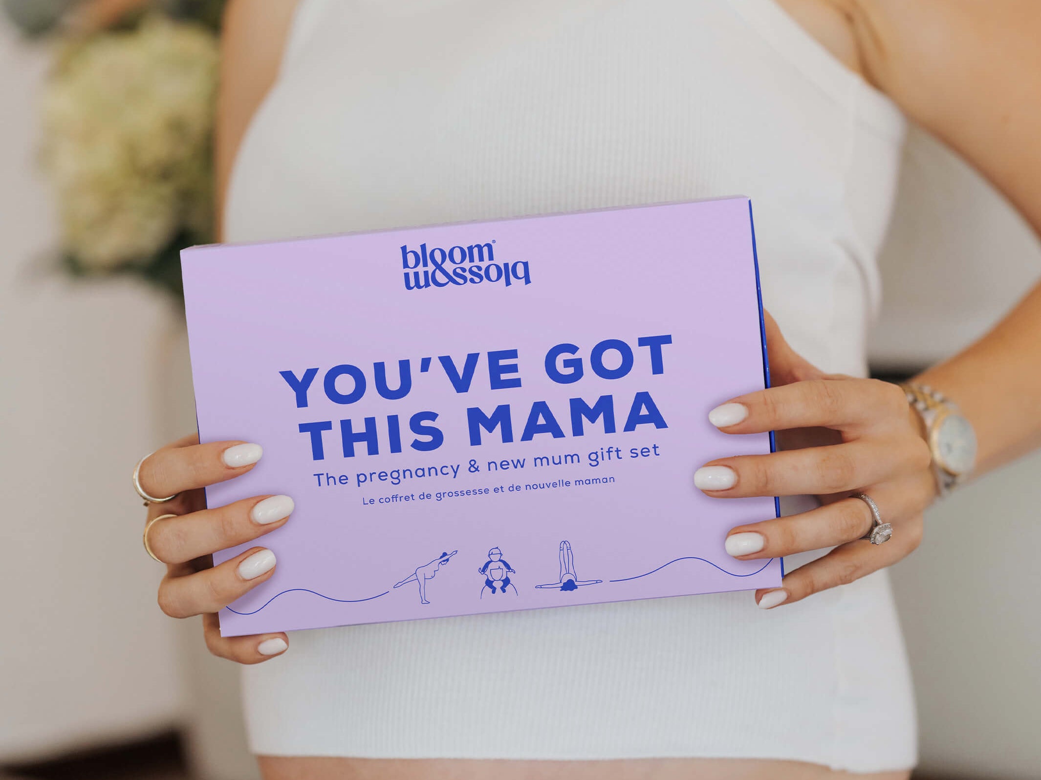 A woman in a white ribbed tank top holds a lavender gift box with blue text that reads 'bloom blossom' and 'YOU'VE GOT THIS MAMA'.