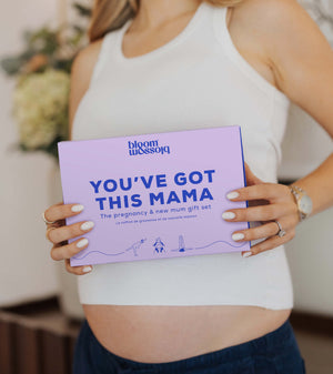 A woman in a white ribbed tank top holds a lavender gift box with blue text that reads 'bloom blossom' and 'YOU'VE GOT THIS MAMA'.