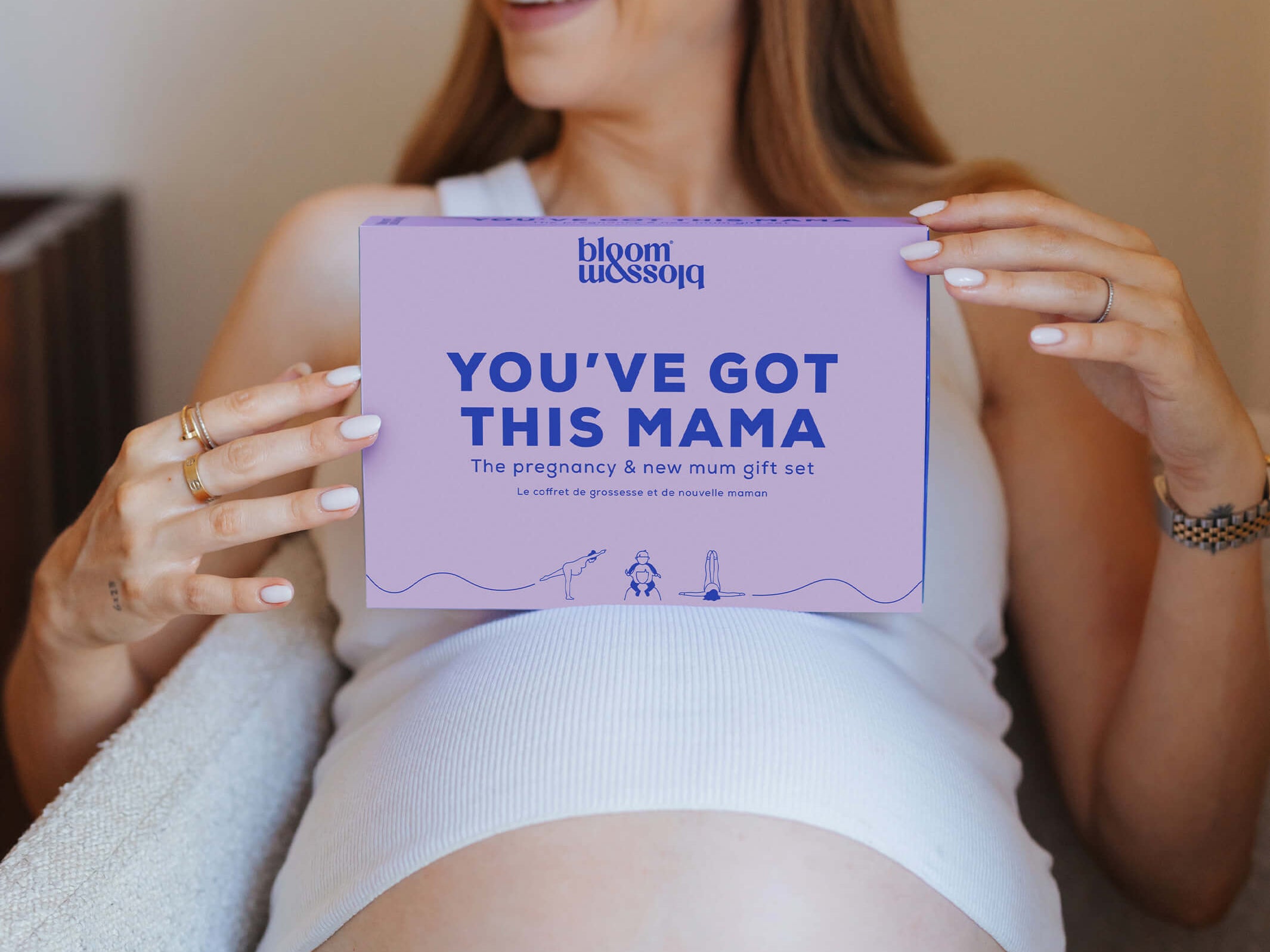 A smiling pregnant woman holds a lavender rectangular card that reads 'YOU'VE GOT THIS MAMA' with the brand name 'bloom with joy' at the top. She is seated indoors against a warm beige wall, wearing a white ribbed tank top and dark navy pants.