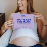 A smiling pregnant woman holds a lavender rectangular card that reads 'YOU'VE GOT THIS MAMA' with the brand name 'bloom with joy' at the top. She is seated indoors against a warm beige wall, wearing a white ribbed tank top and dark navy pants.