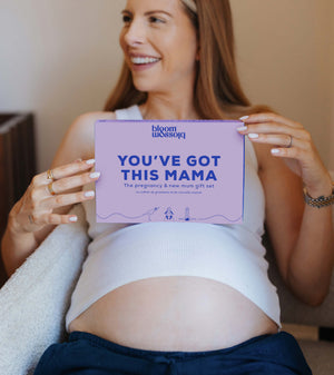 A smiling pregnant woman holds a lavender rectangular card that reads 'YOU'VE GOT THIS MAMA' with the brand name 'bloom with joy' at the top. She is seated indoors against a warm beige wall, wearing a white ribbed tank top and dark navy pants.