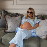 A pregnant woman wearing a light blue maternity sleep set and sunglasses reclines on a green couch with patterned pillows.