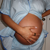 A close-up of a pregnant person wearing a light blue and white gingham maternity top and matching pants, holding their baby bump with one hand while wearing rings with the other.