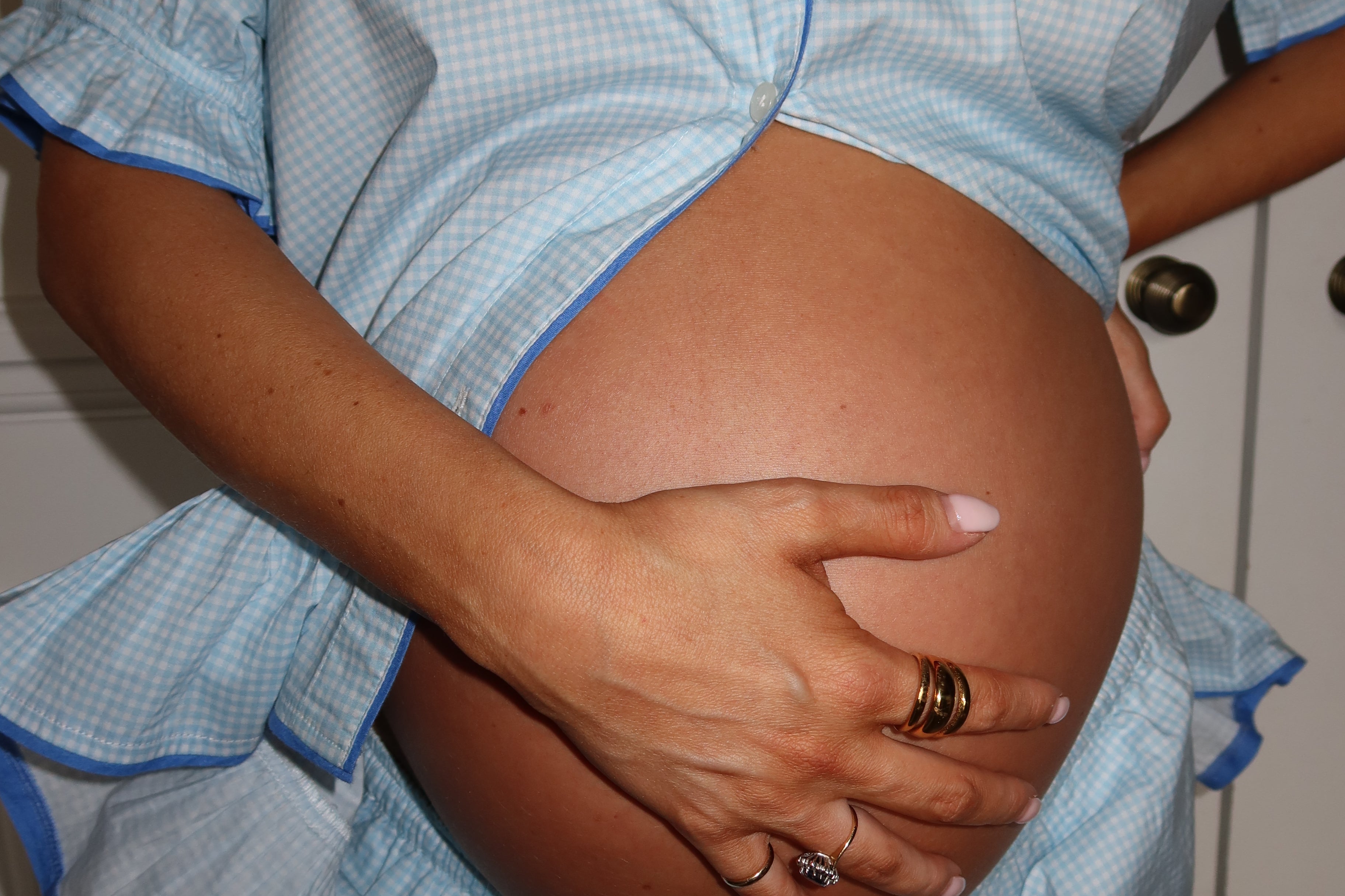 A close-up of a pregnant person wearing a light blue and white gingham maternity top and matching pants, holding their baby bump with one hand while wearing rings with the other.