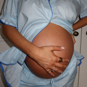 A close-up of a pregnant person wearing a light blue and white gingham maternity top and matching pants, holding their baby bump with one hand while wearing rings with the other.