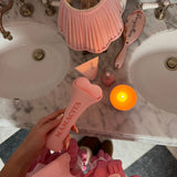 A person in a pink gingham dress holds a pink Mamacita hair straightener over a marble bathroom counter with a pink pleated lamp, candle, and hairbrush nearby.