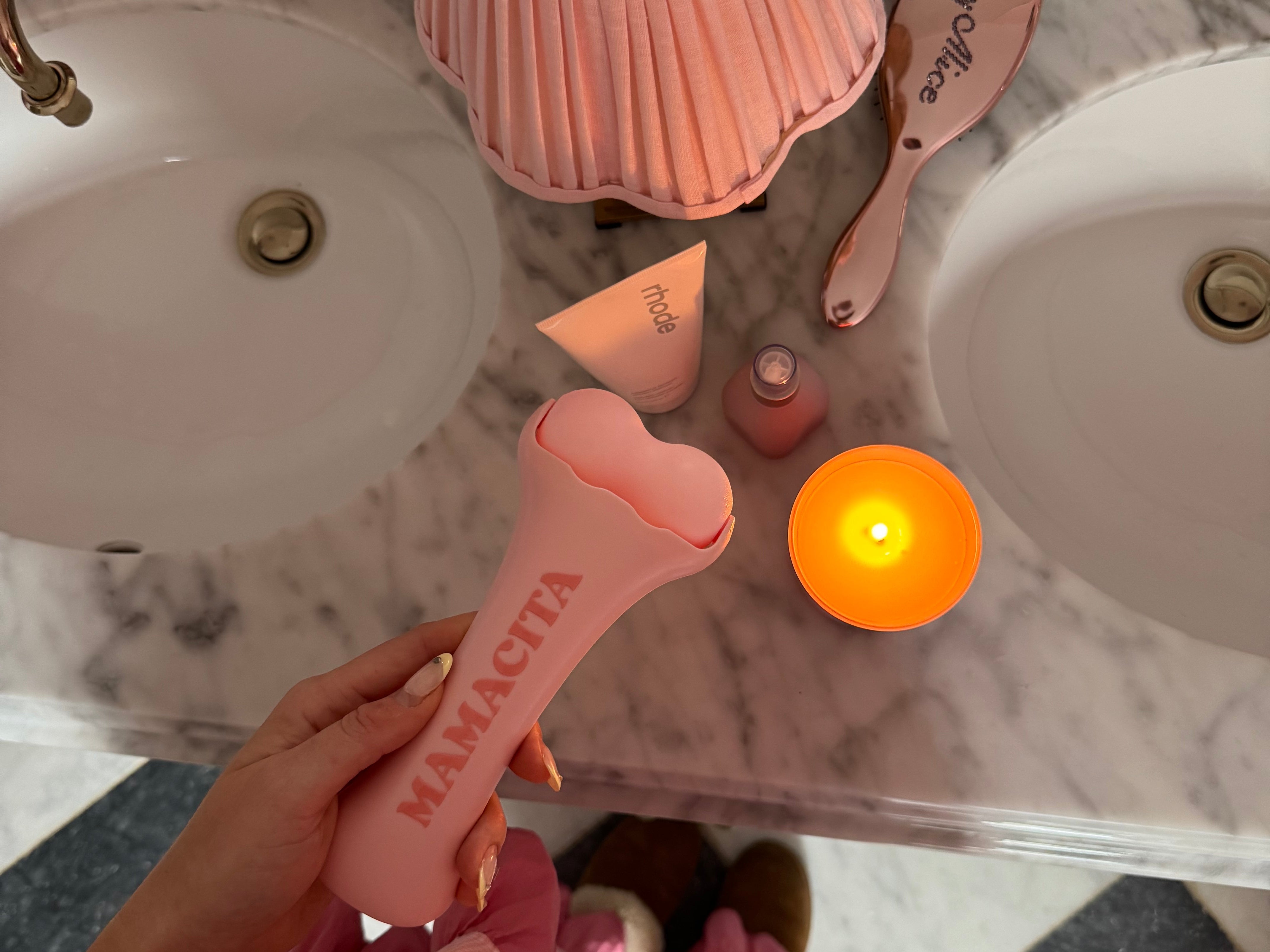 A person in a pink gingham dress holds a pink Mamacita hair straightener over a marble bathroom counter with a pink pleated lamp, candle, and hairbrush nearby.