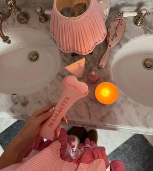 A person in a pink gingham dress holds a pink Mamacita hair straightener over a marble bathroom counter with a pink pleated lamp, candle, and hairbrush nearby.