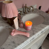 A pink facial massage tool with the brand name 'MAMAGITA' printed on it, styled on a marble bathroom vanity alongside a pink pleated lamp, a pink candle, and other toiletries.