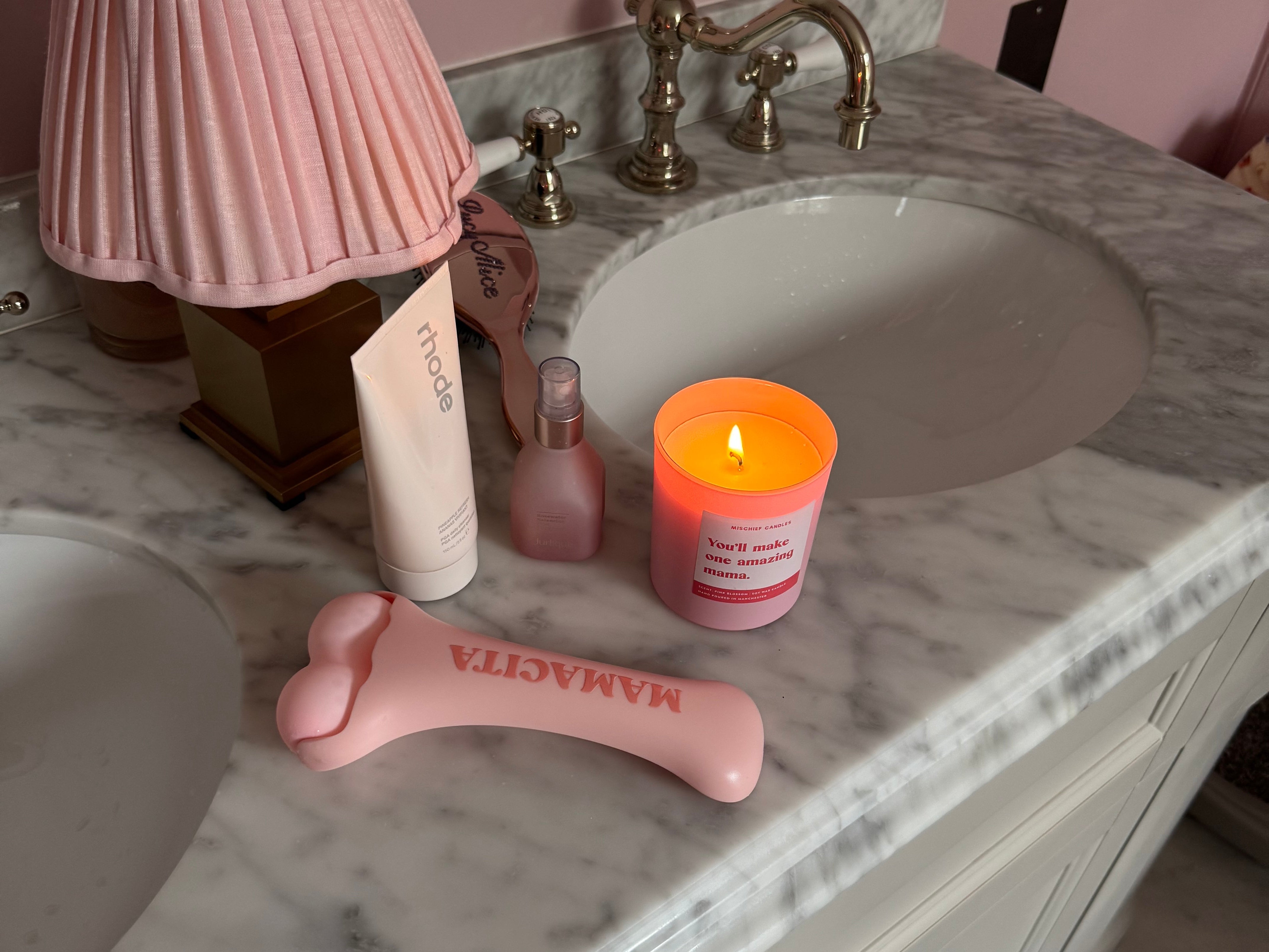 A pink facial massage tool with the brand name 'MAMAGITA' printed on it, styled on a marble bathroom vanity alongside a pink pleated lamp, a pink candle, and other toiletries.