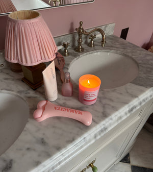 A pink facial massage tool with the brand name 'MAMAGITA' printed on it, styled on a marble bathroom vanity alongside a pink pleated lamp, a pink candle, and other toiletries.
