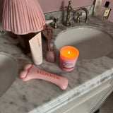 A pink silicone prostate massager lies on a white marble bathroom vanity next to a pink pleated lamp, a pink candle, and other toiletries.